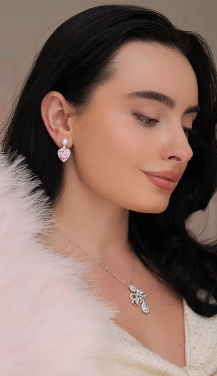 A silver-toned drop earring with a white pearl stud, suspending a pink crystal heart framed by clear crystals.