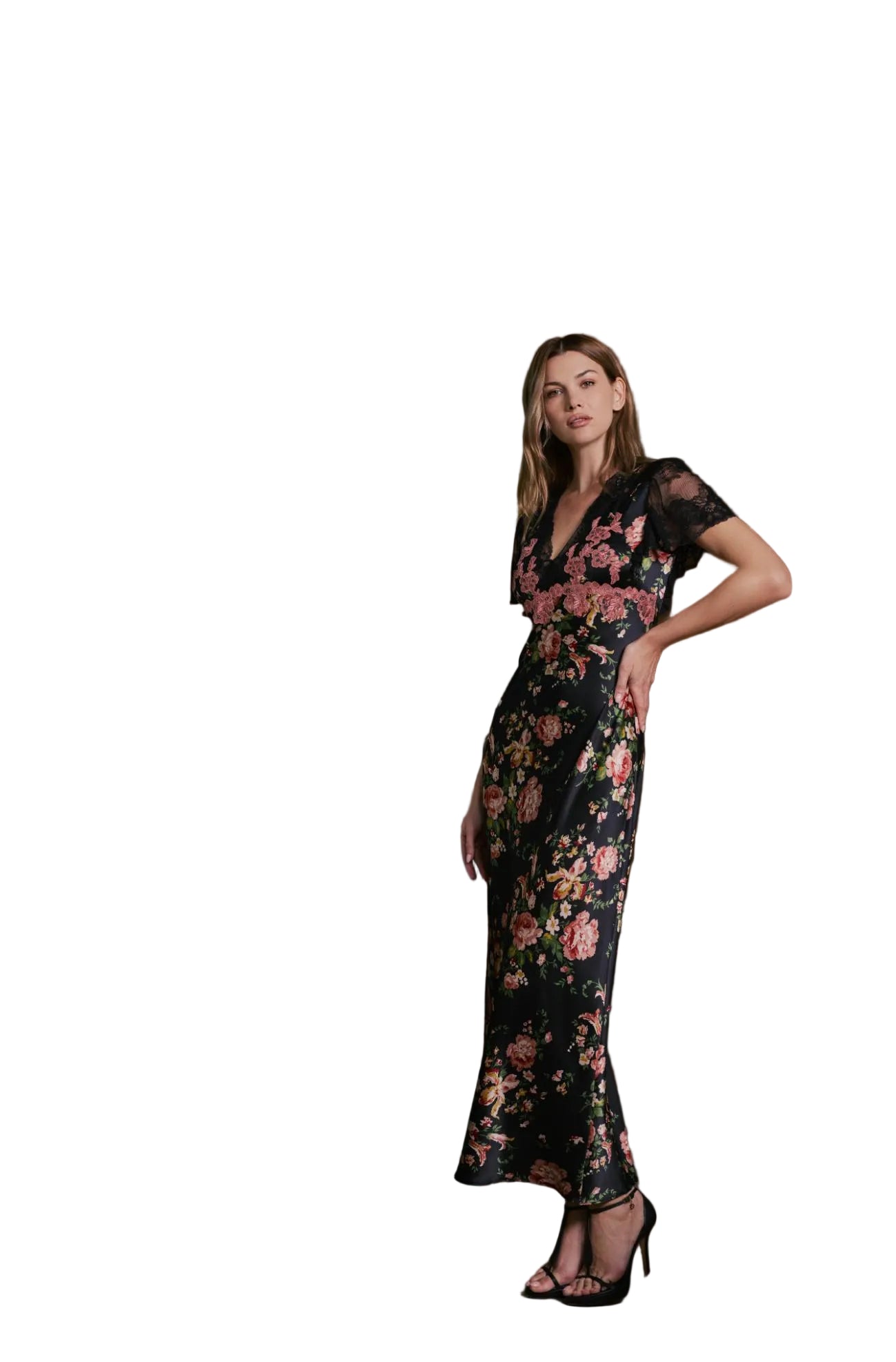 A woman models a black floral maxi dress with sheer short sleeves and a V-neckline, standing in a modern interior setting with concrete stairs and a wooden slatted wall.