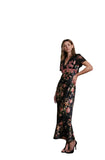 A woman models a black floral maxi dress with sheer short sleeves and a V-neckline, standing in a modern interior setting with concrete stairs and a wooden slatted wall.