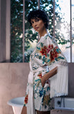 A woman wearing a white silk-like robe with a colorful floral print and long fringe on the sleeves, standing in a bathroom with a bathtub and large windows in the background.