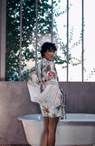 A woman stands in front of a white freestanding bathtub in a modern interior with tall vertical bars and greenery visible behind her, wearing a white fringed kimono-style cover-up with a colorful floral print and a matching floral mini skirt.