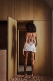 A woman stands with her back to the camera in a doorway, wearing a white fringed lingerie-style dress with strappy back detailing. The setting features warm wood-paneled walls and a patterned rug.