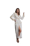 A woman wearing a white satin robe with gold embroidery and a matching belt stands in a modern architectural setting with concrete steps and wooden slats.