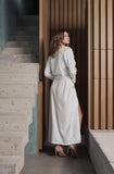 A woman wearing a long white wrap-style dress with a high neck and side slit, standing in a modern interior with concrete stairs and wood slat walls.