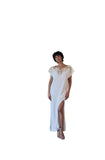 A woman stands in front of a large concrete fireplace wearing a long white slip dress with gold embroidery on the bodice and a high slit.