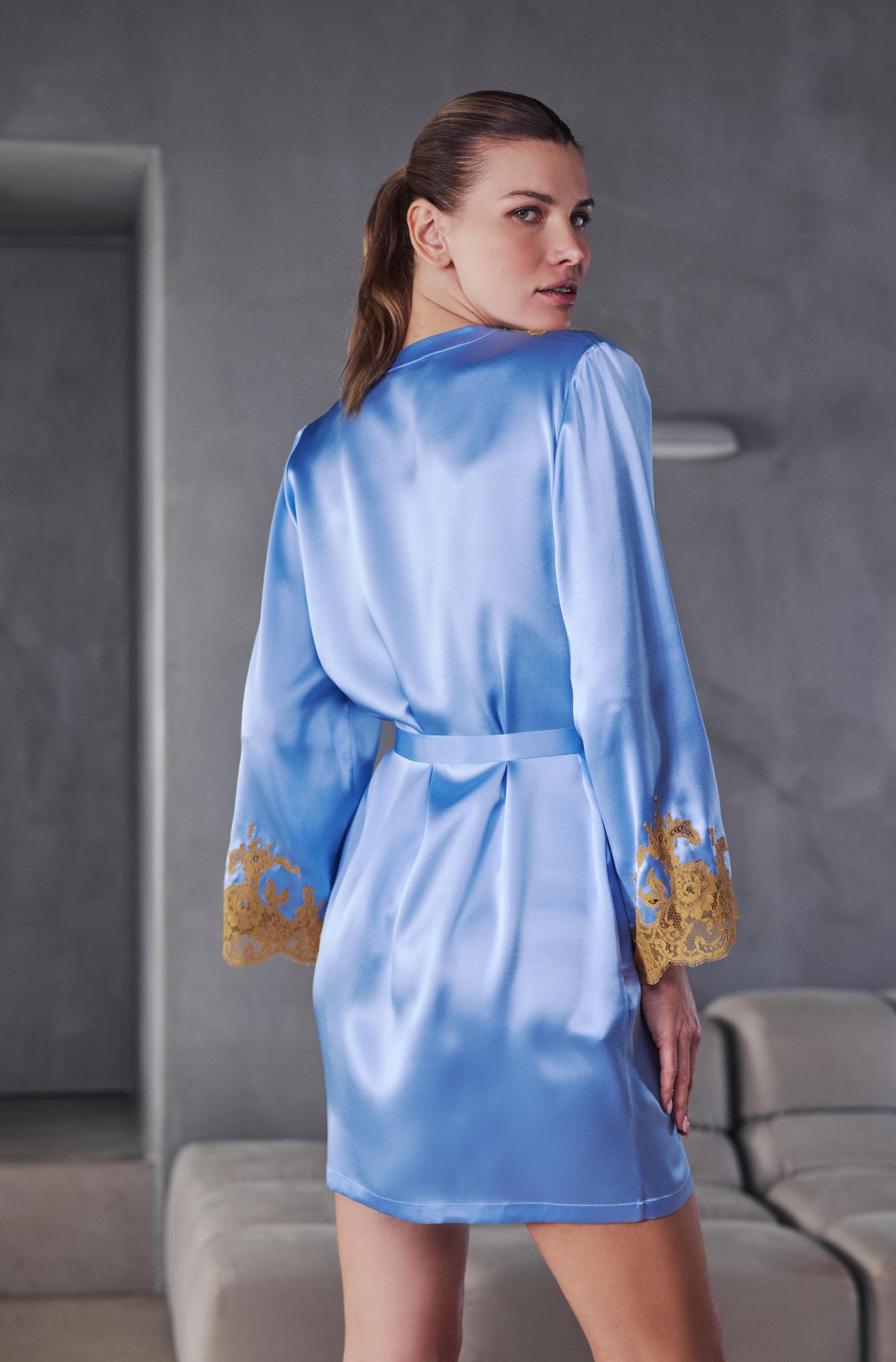 A woman wearing a light blue satin robe with gold embroidered cuffs, shown from the back in a modern gray interior setting.