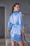 A woman wearing a light blue satin robe with gold embroidered cuffs, shown from the back in a modern gray interior setting.