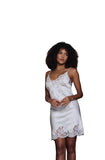 A woman with curly dark hair models a white satin slip dress with lace trim, standing in a modern interior setting with a teal wall, concrete stairs, and a slatted wood door.