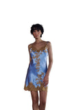 A woman wearing a light blue satin slip dress with gold floral embroidery and lace trim, standing in a doorway with a gray wall and terracotta-colored stairs behind her.