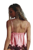 A woman stands on a balcony wearing a pink satin lingerie set with black lace trim, photographed from behind.