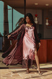 A woman wearing a pink satin slip dress with dark floral lace trim and a matching sheer dark plum robe, standing in a modern interior setting.