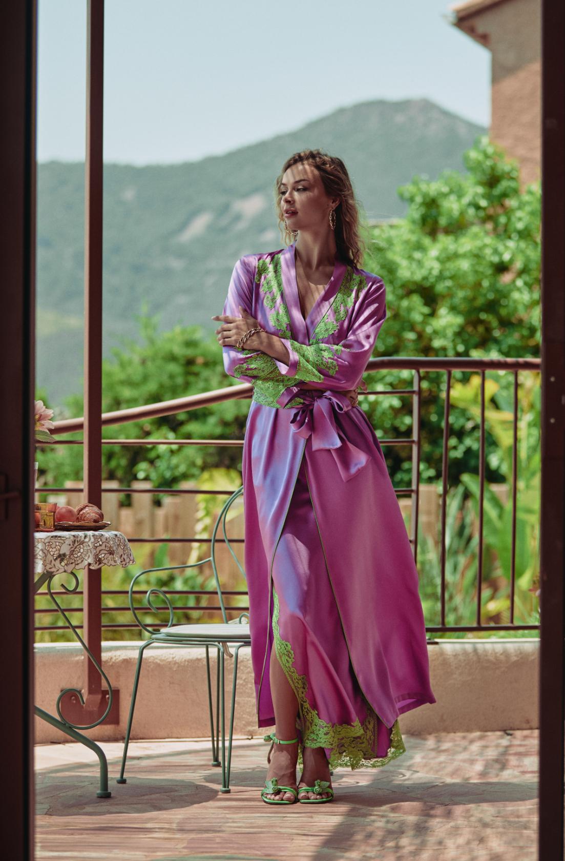 A woman stands on a balcony wearing a long purple satin robe with green lace trim and matching green sandals, with a mountain and garden visible in the background.