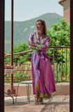 A woman stands on a balcony wearing a long purple satin robe with green lace trim and matching green sandals, with a mountain and garden visible in the background.