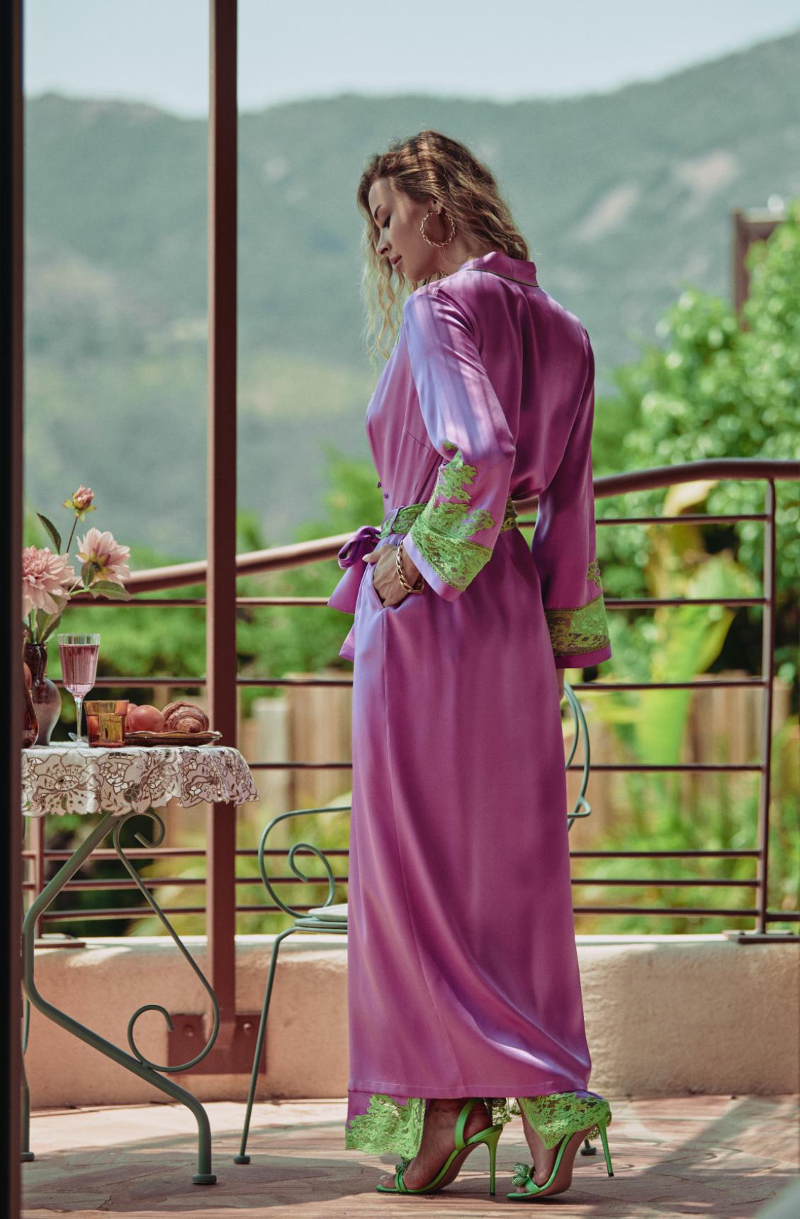 A woman stands on a balcony wearing a long magenta-purple robe with green patterned trim and green high heels, with a table and outdoor greenery in the background.