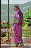 A woman stands on a balcony wearing a long magenta-purple robe with green patterned trim and green high heels, with a table and outdoor greenery in the background.