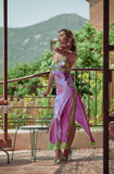 A woman wearing a flowing lavender and purple maxi dress with green lace trim stands on a sunlit outdoor patio with a mountain and greenery in the background.