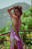 A woman posing on a balcony wearing a purple satin slip dress with green floral embroidery and lace-up back details, with a blurred green garden background.