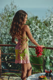 A woman stands on a balcony wearing a short, colorful dress with pink, green, and yellow floral patterns and crisscross back straps. She is holding a bouquet of red flowers, with a table and green plants in the background.