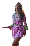 A woman wearing a purple silk-like robe with green floral embroidery and a matching tie belt, standing on an outdoor balcony with a blurred mountain background.