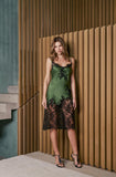 A woman models a green satin slip dress with black floral lace detailing, standing in front of a modern wooden slatted wall.