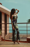 A woman stands on a wooden deck wearing a sheer black lace slip dress with a colorful floral print, posing outdoors with a beach and ocean in the background.