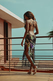 Woman in a floral dress standing on a balcony with a clear blue sky and ocean view.