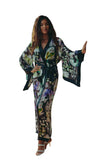 A woman models a long-sleeved, floor-length floral print robe with a tie belt, standing on a stone patio in front of a glass door.