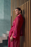 A woman in profile wearing a sheer magenta-pink patterned robe, leaning against a modern wooden slatted wall next to a staircase.