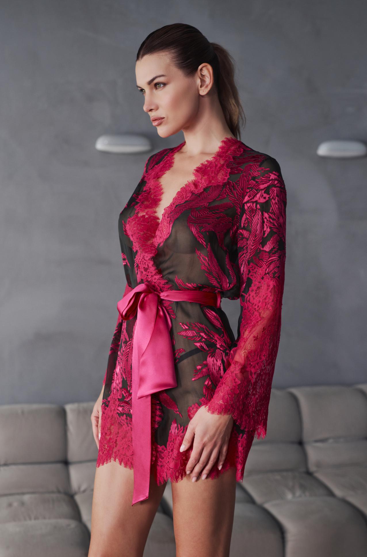 A woman models a sheer black lace robe with vibrant magenta floral patterns and a matching magenta satin tie belt, set against a gray textured wall and sofa background.