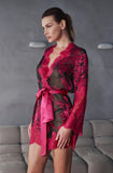 A woman models a sheer black lace robe with vibrant magenta floral patterns and a matching magenta satin tie belt, set against a gray textured wall and sofa background.