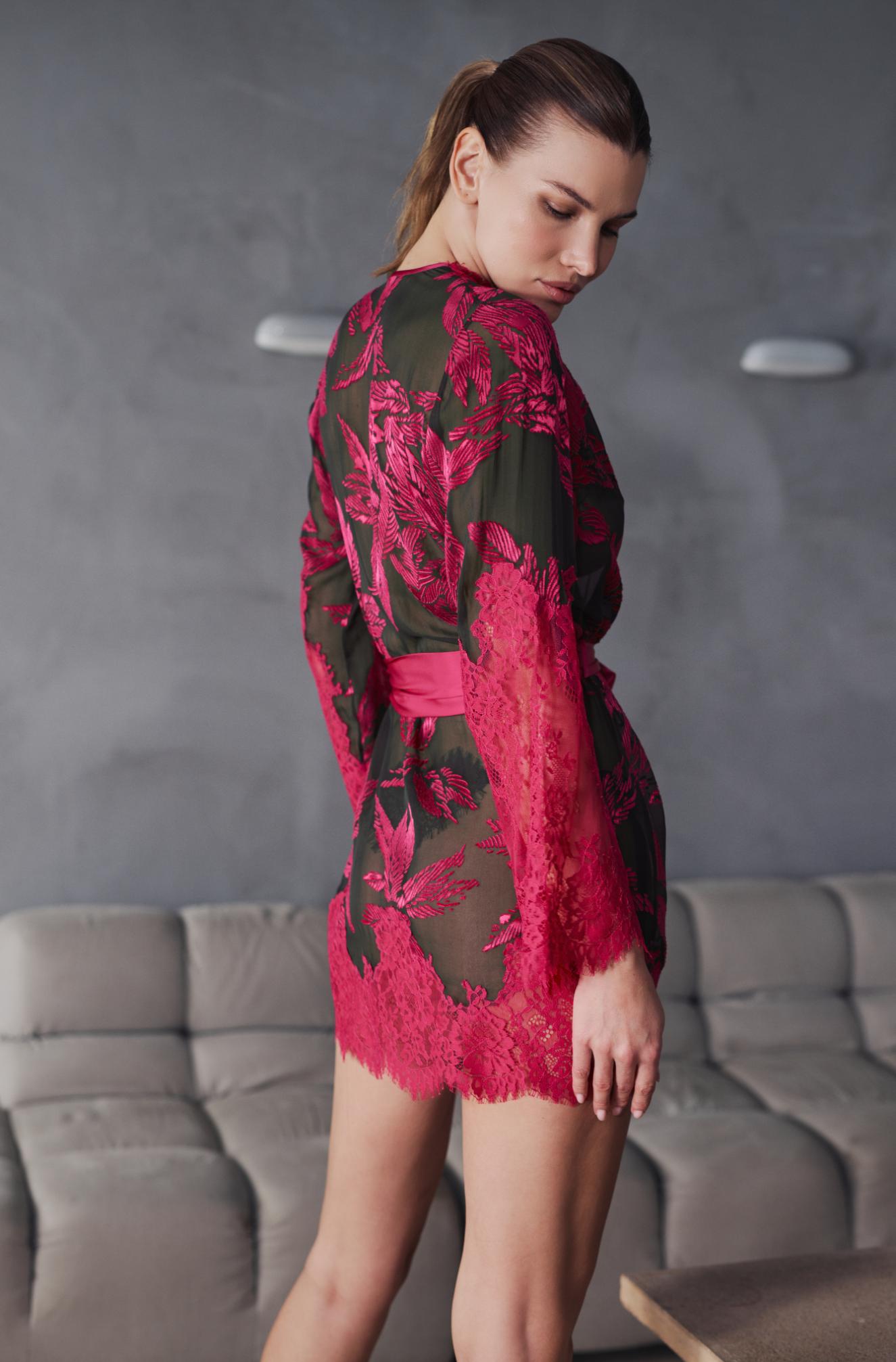 A woman wearing a sheer dark green robe-style mini dress with vibrant pink floral lace patterns and a matching pink waist tie, standing in a modern room with a gray tufted sofa.