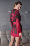 A woman wearing a sheer dark green robe-style mini dress with vibrant pink floral lace patterns and a matching pink waist tie, standing in a modern room with a gray tufted sofa.