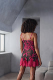 A woman viewed from behind wearing a sheer black slip dress with a vibrant pink floral lace overlay and thin pink straps, standing in a minimalist gray room.