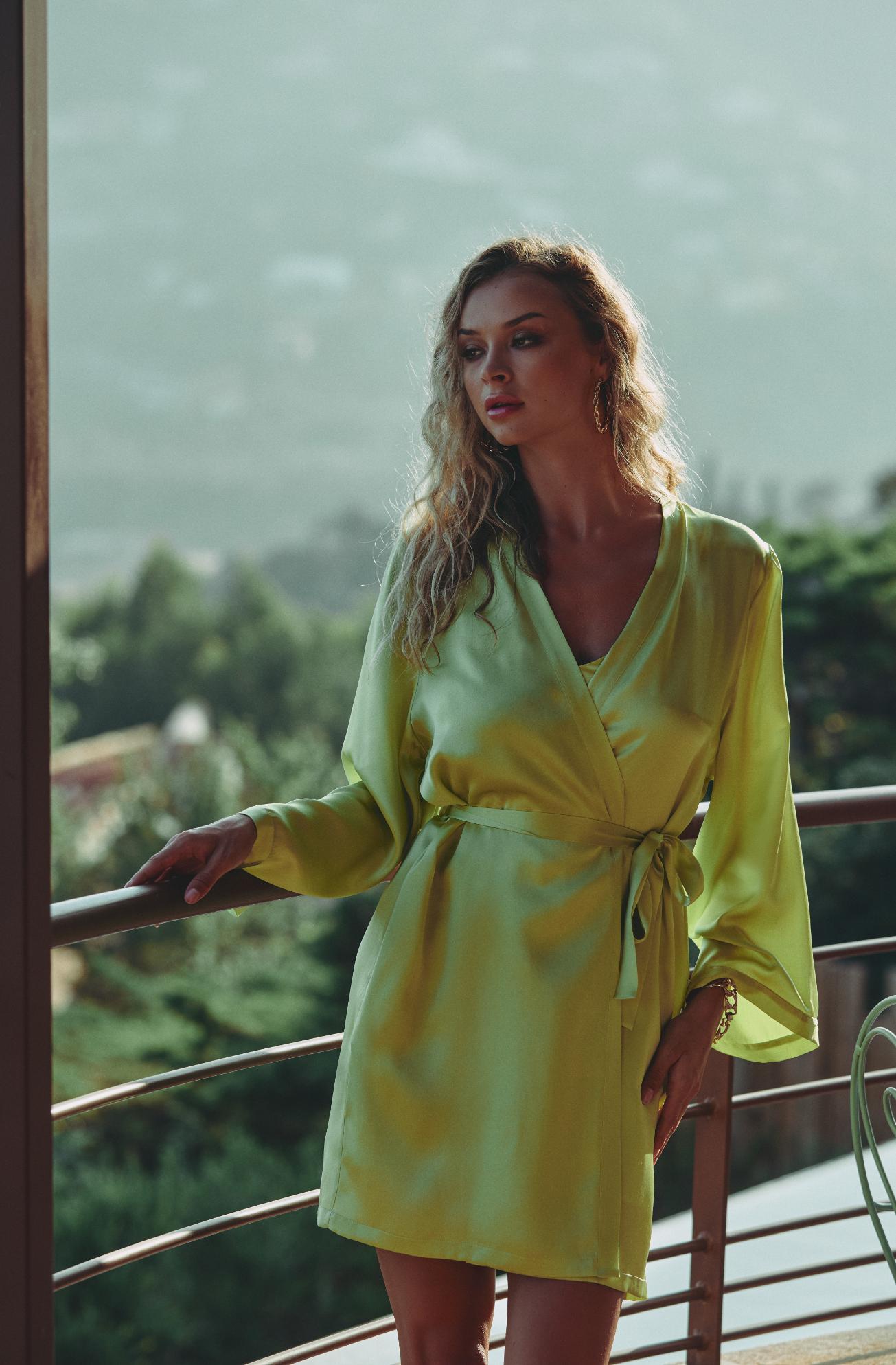 A woman wearing a lime green satin wrap robe stands on a balcony with a scenic outdoor view in the background.