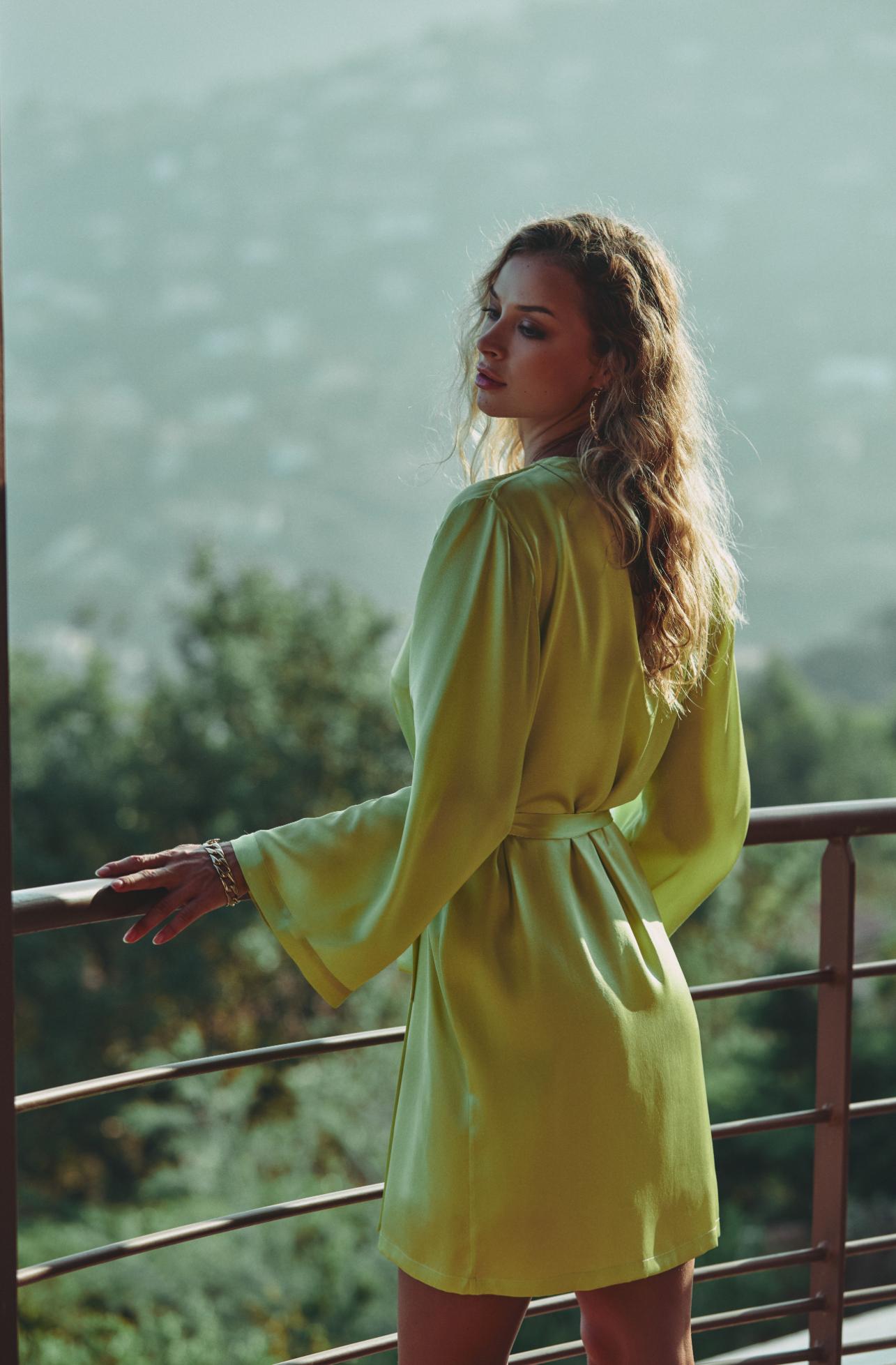 A woman wearing a bright yellow-green long-sleeve mini dress with a tie waist, standing on a balcony with a scenic, blurred mountain background.