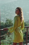 A woman wearing a bright yellow-green long-sleeve mini dress with a tie waist, standing on a balcony with a scenic, blurred mountain background.