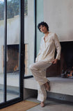 A woman with short curly dark hair wearing a cream-colored linen pajama set consisting of a long-sleeve button-up shirt and matching wide-leg pants, sitting on a concrete ledge next to a large glass window in a modern minimalist room.