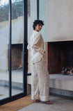 A model wearing a light beige linen two-piece set consisting of a long-sleeve button-up shirt and matching wide-leg trousers, standing in a modern minimalist room with a large window and fireplace.