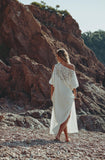 A woman wearing a white one-shoulder bohemian maxi dress with lace embroidery on the back, walking barefoot on a rocky beach with a large reddish-brown cliff in the background.