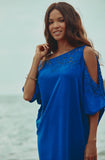 A woman wearing a bright blue cold-shoulder kaftan dress with lace detailing, standing outdoors against a soft-focus beach or water background.