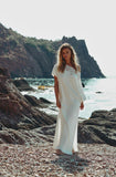 Woman in a white dress standing on a rocky beach with cliffs in the background.
