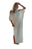 A woman wearing a white off-the-shoulder maxi dress with lace detailing and a thigh-high slit, standing on a pebble beach with rocks and the ocean in the background.