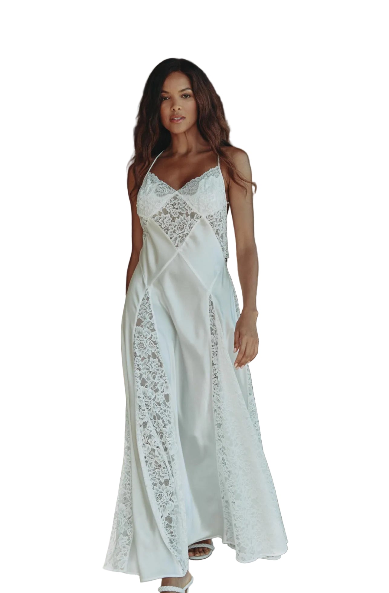 A woman wearing a white satin slip dress with intricate lace panels, standing in a warmly lit, rustic living room with brown leather furniture and a green spiral floor lamp.