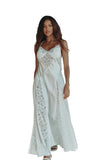 A woman wearing a white satin slip dress with intricate lace panels, standing in a warmly lit, rustic living room with brown leather furniture and a green spiral floor lamp.