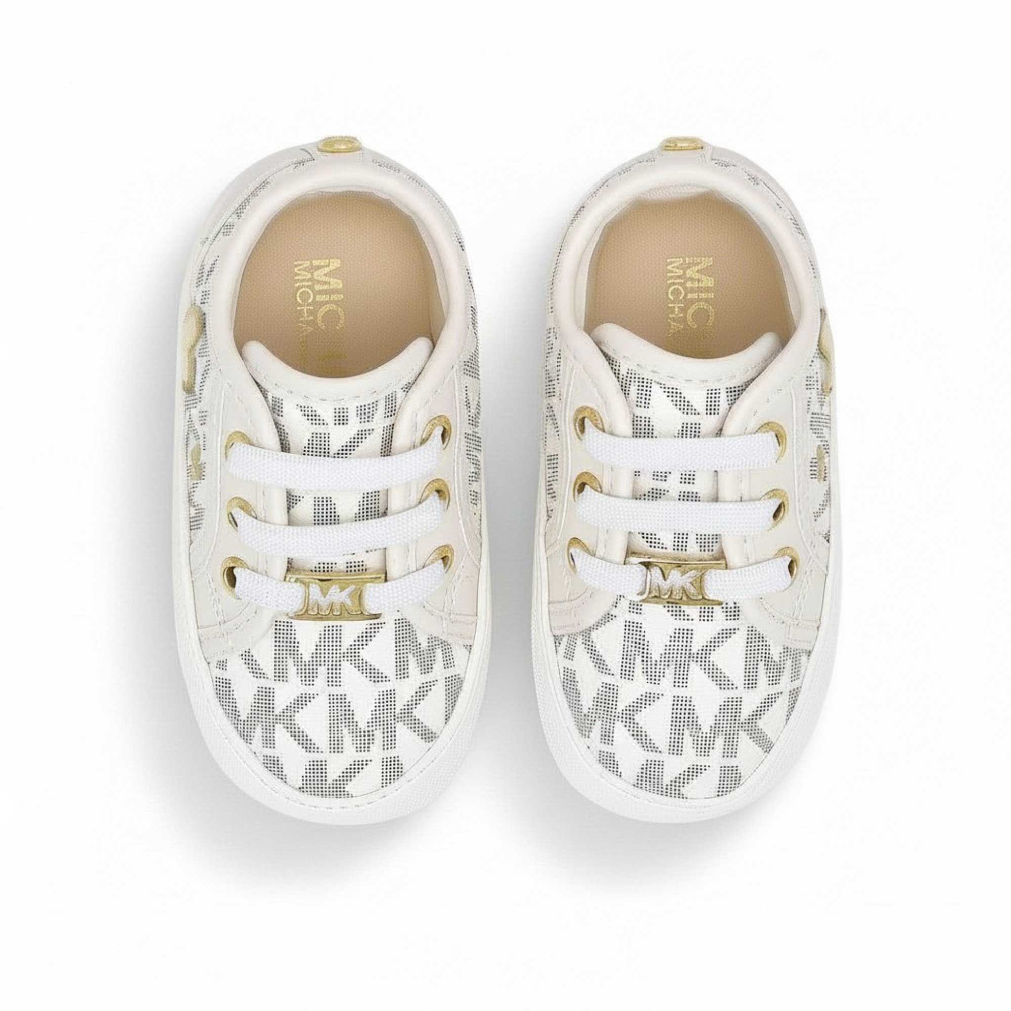 Front view of a pair of off-white Michael Kors infant shoes, showing white laces, gold metallic eyelets, and a shiny gold-tone MK monogram lace lock hardware piece.