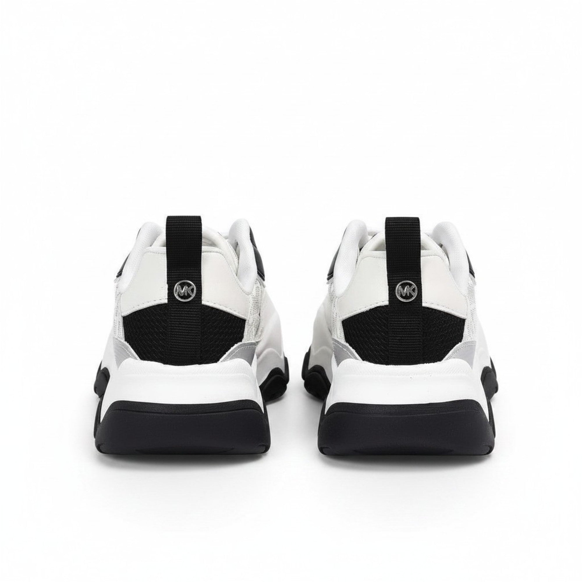 Rear heel view of a pair of kids' black and white Michael Kors lifestyle runners, showing white upper panels, black webbed heel loops, and small circular silver MK metallic badges.