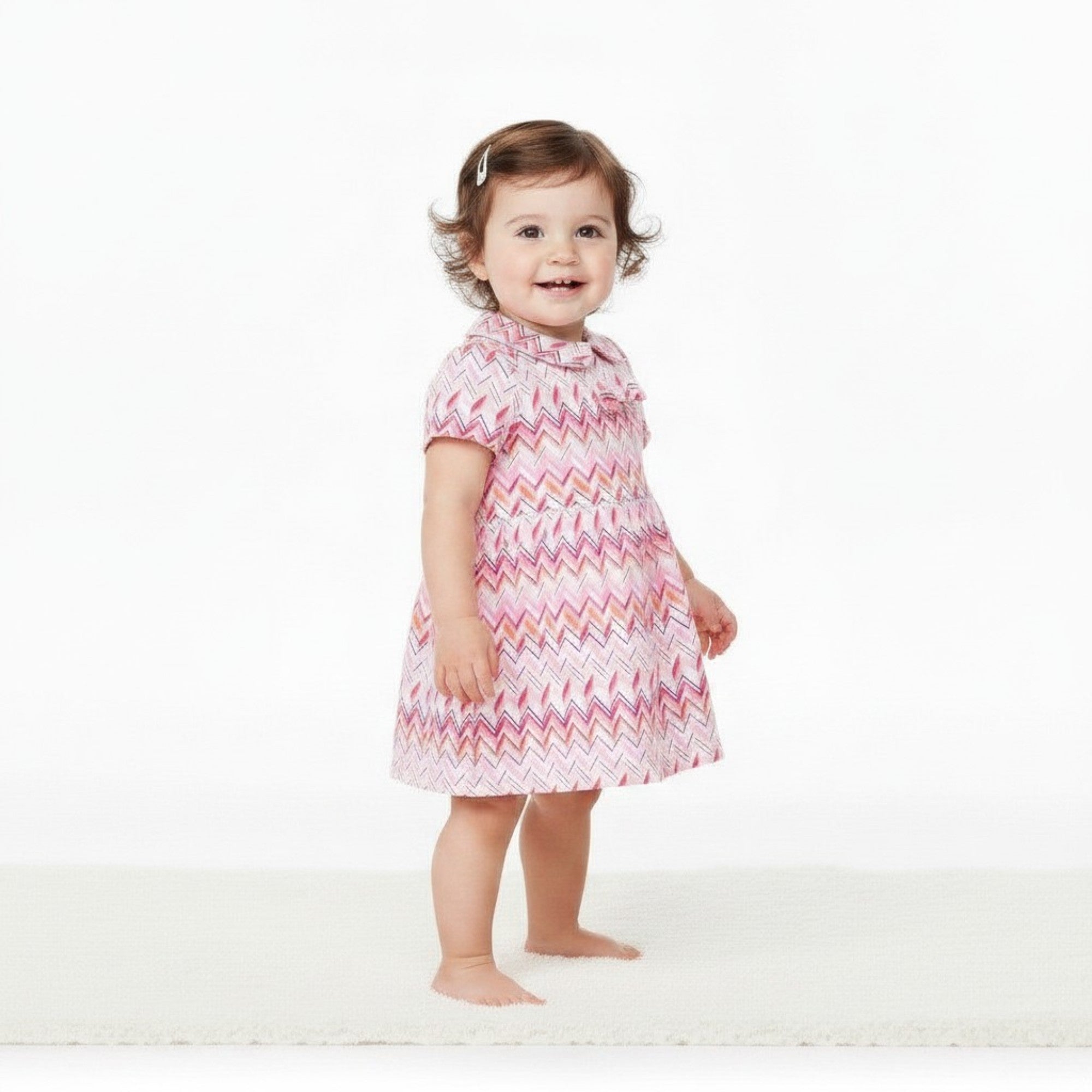 A smiling baby girl model presenting a full-length side view of the pink and orange Missoni chevron dress with a Peter Pan collar against a white backdrop.