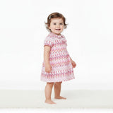 A smiling baby girl model presenting a full-length side view of the pink and orange Missoni chevron dress with a Peter Pan collar against a white backdrop.
