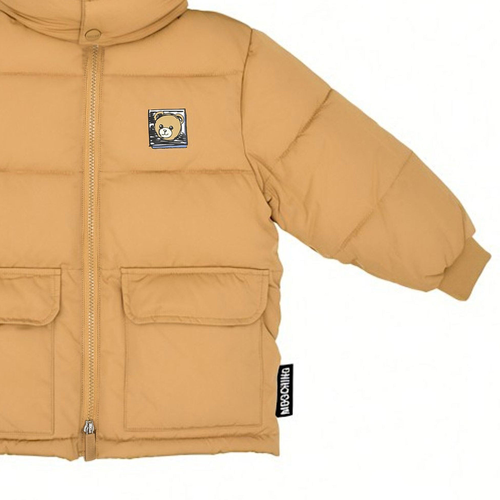 Close-up cropped image focusing on the right side of the Moschino Kids camel jacket, displaying a large front flap pocket, ribbed cuff, zip line, and the vertical brand tag.