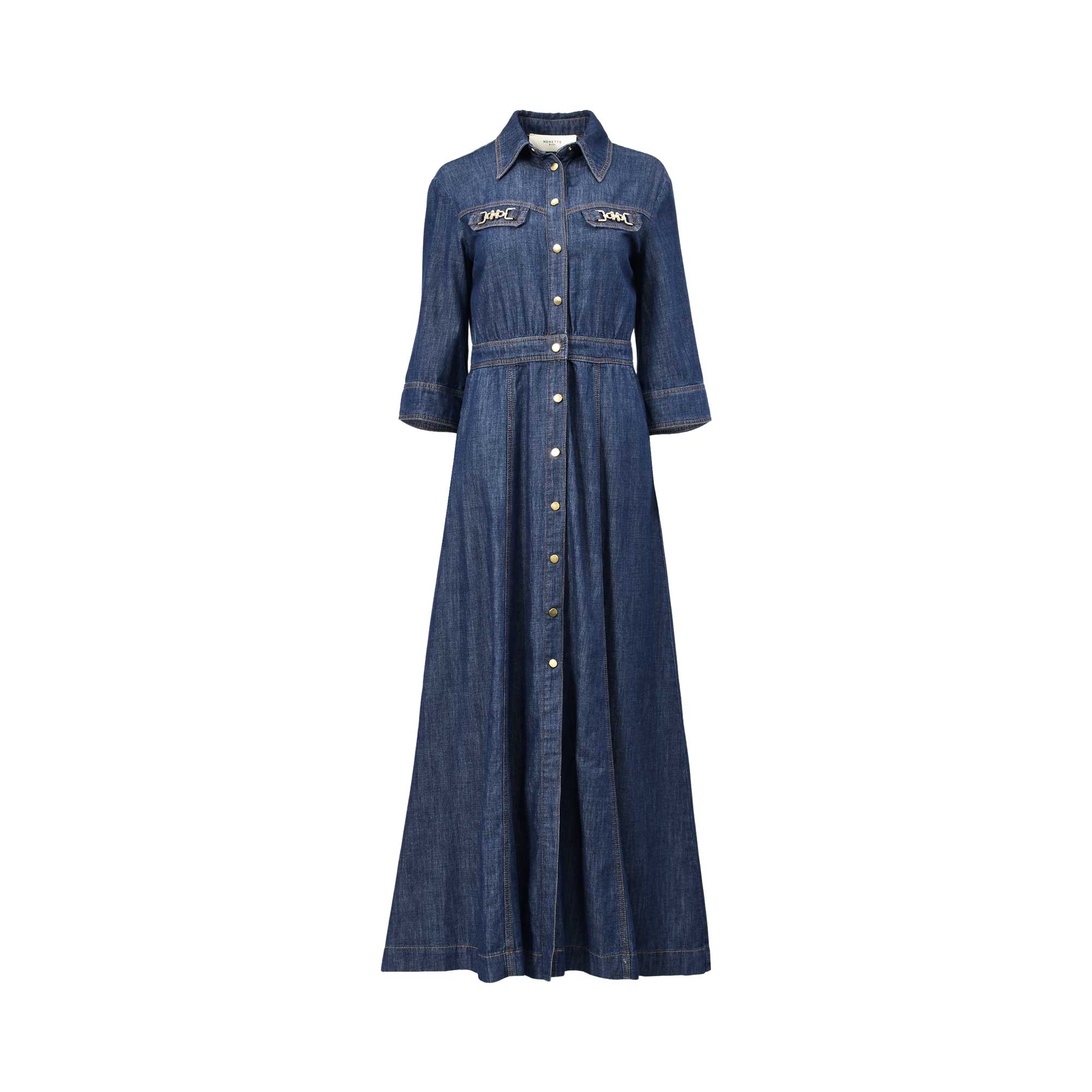 Front studio view of a dark blue denim maxi shirt dress by Nenette against a plain white background, highlighting its button-down front, pointed collar, and metallic chain accents on the chest.